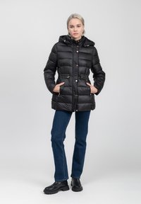 Zwarte puffy parka met capuchon, voorzijde met ritssluiting en zijzakken. Voorzien van gouden accenten en een getailleerde taille. Gecombineerd met blauwe jeans en zwarte laarzen.