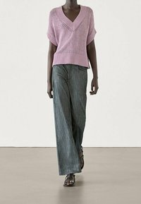 Laventelinsinis megztinis trumpomis rankovėmis su V formos iškirpte, derinamas su plačiomis pilkomis kelnių ir juodais sandalais, rodomas neutraliame fone.