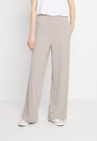 Beżowe spodnie o szerokich nogawkach z elastycznym paskiem w talii i bocznymi kieszeniami, wykonane z gładkiego, lekkiego materiału. Nosi się je z białymi trampkami.