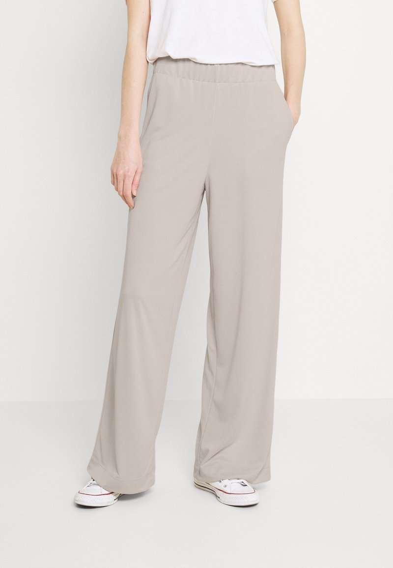 Beżowe spodnie o szerokich nogawkach z elastycznym paskiem w talii i bocznymi kieszeniami, wykonane z gładkiego, lekkiego materiału. Nosi się je z białymi trampkami.