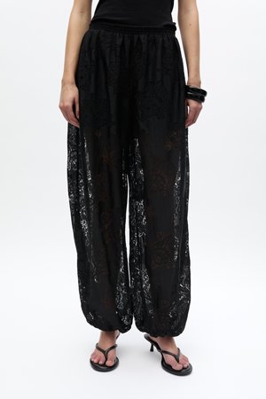 Femme portant un pantalon large en dentelle noire transparente avec des poignets élastiques, un top noir sans manches, un bracelet noir et des sandales à talons noires.