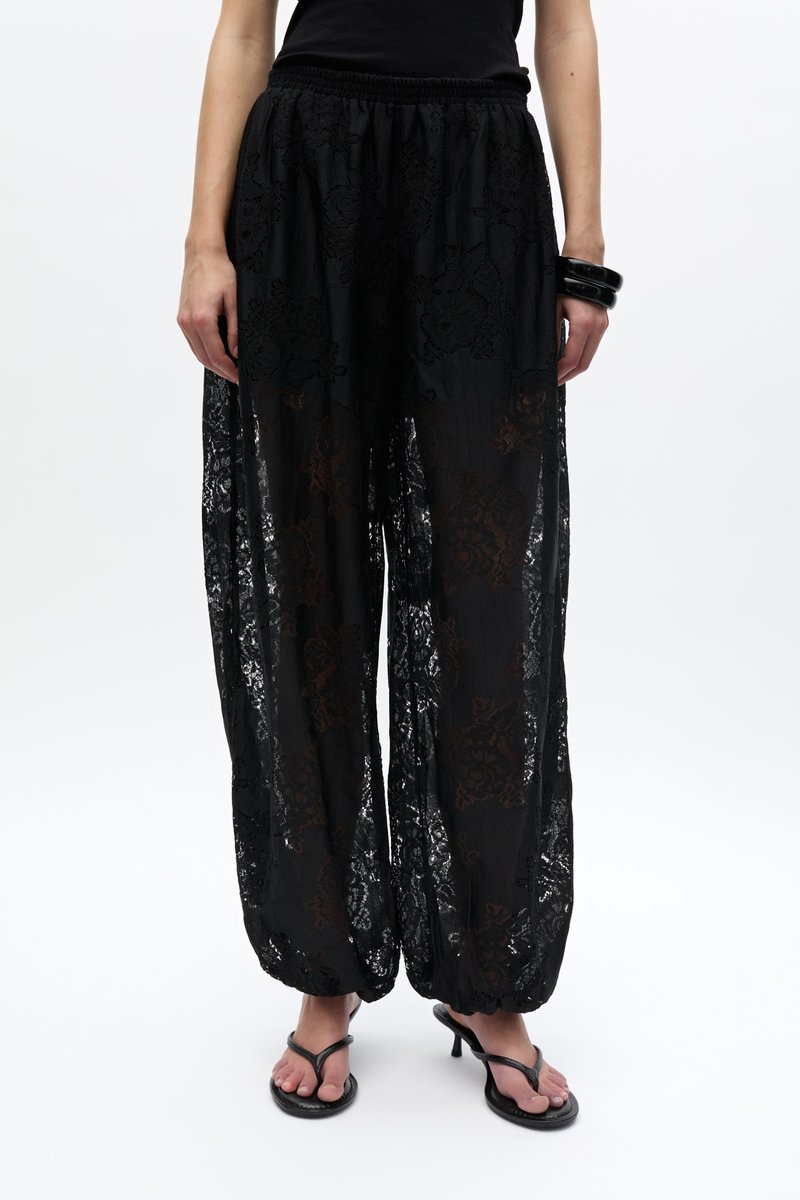 Femme portant un pantalon large en dentelle noire transparente avec des poignets élastiques, un top noir sans manches, un bracelet noir et des sandales à talons noires.