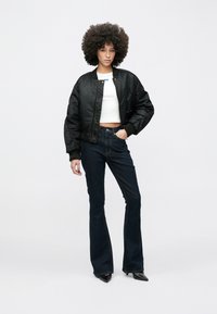 Tânără cu coafură afro, purtând o jachetă bomber neagră, top scurt alb, blugi evazați închiși la culoare și pantofi negri cu toc ascuțit, pe un fundal alb.