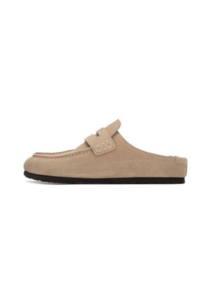 Zapato beige de ante tipo slip-on con punta redondeada, correa decorativa con hebilla y suela negra texturizada. Superficie lisa, detalle de costuras mínimas.