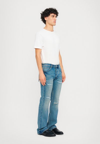 Ελαφρύ λευκό βαμβακερό μπλουζάκι συνδυασμένο με μπλε τζιν παντελόνι που διαθέτει φθαρμένες λεπτομέρειες, φαρδιά πόδια και μπροστινές τσέπες. Μαύρα παπούτσια ολοκληρώνουν την εμφάνιση.