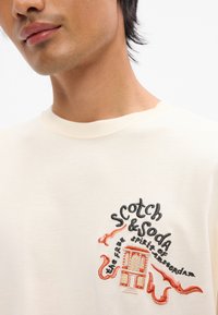 Μπεζ βαμβακερό t-shirt με περίπλοκο κεντημένο λογότυπο σε μαύρο και πορτοκαλί, που απεικονίζει ένα κτίριο και στυλιζαρισμένο κείμενο. Στρογγυλή λαιμόκοψη.