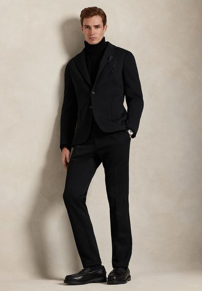 Μαύρο κοστούμι ραμμένο κατά παραγγελία με εφαρμοστό σακάκι, ζιβάγκο και στενά παντελόνια, σετ με μαύρα δερμάτινα παπούτσια. Διακριτικό υφασμάτινο υφή και κομψές καρφίτσες.