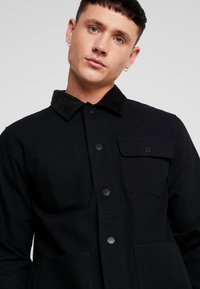 Black jacket with a corduroy collar, two chest pockets, button closure, and a smooth texture. Functional design with a tailored fit.