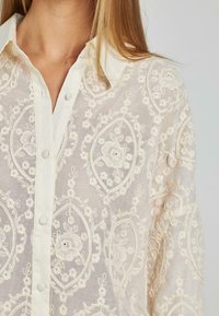 Bestickte Bluse in hellem Beige mit floralem Muster. Verfügt über einen Kragen und einen Knopfverschluss, besteht aus durchscheinendem Stoff mit filigranen Details.