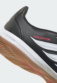 Chaussure de football en salle noire avec des détails à rayures blanches, texte rouge "PREDATOR", semelle intermédiaire blanche et semelle extérieure en gomme texturée.