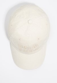 Gorra de pana en color crema con un logo bordado. Presenta costillas elevadas, una corona redondeada y una visera curva. Diseño simple y texturizado.