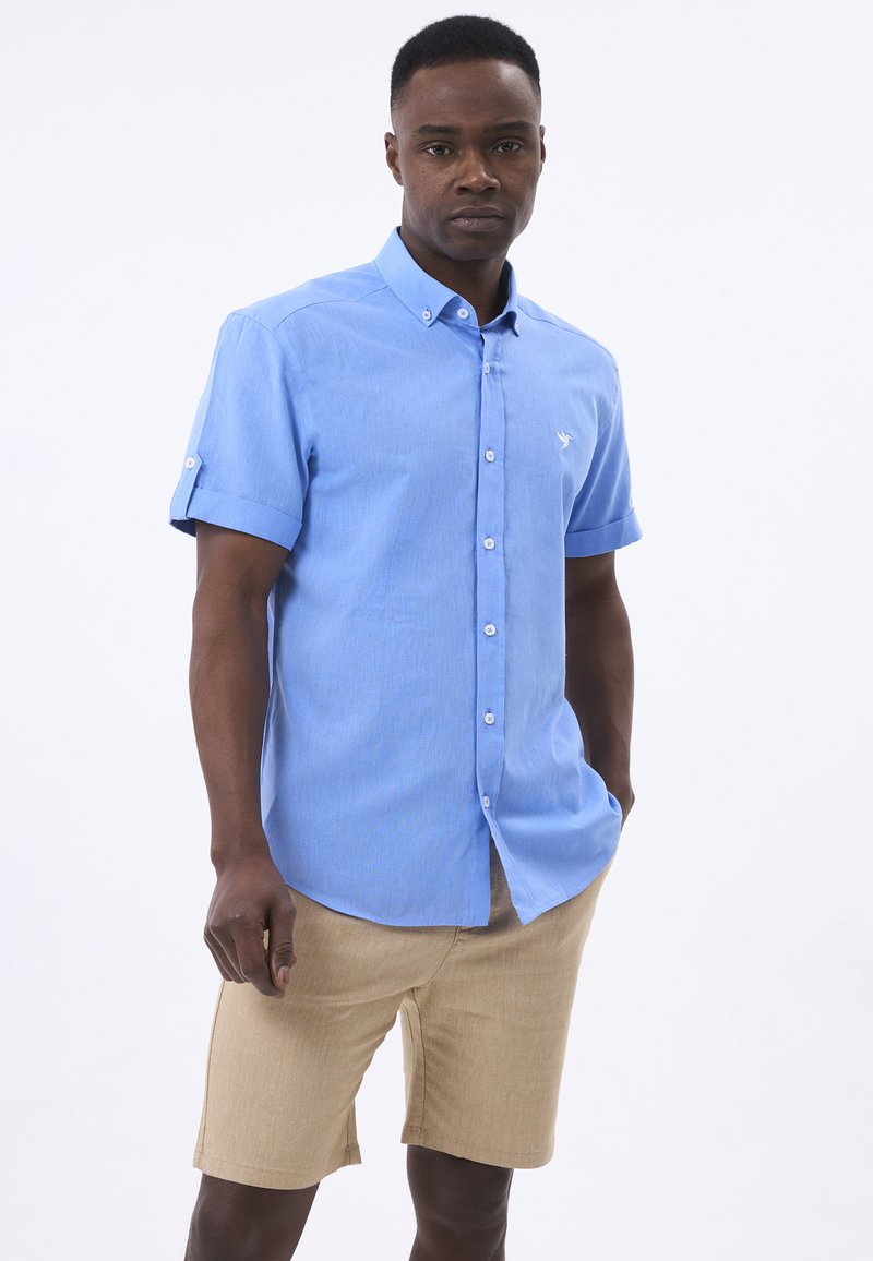 Himmelblaues kurzärmliges Button-Up-Hemd aus strukturiertem Stoff, mit einem kleinen Logo auf der Brust. Kombiniert mit beigen Shorts.