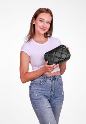 Schwarze, gesteppte Handtasche mit strukturiertem Finish und Reißverschluss, gehalten von einer Person, die ein weißes, enges Oberteil und blaue Jeans trägt.