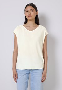 Top giallo chiaro a maniche corte con scollatura a V e texture morbida e liscia. Indossato con jeans in denim blu chiaro, che mostrano un fit casual.