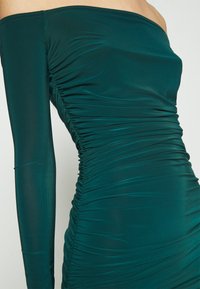 Robe longue bleu sarcelle épaules dénudées avec des détails froncés sur les côtés et une silhouette ajustée.