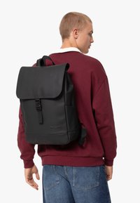 Sac à dos noir en matériau mat, avec un rabat de fermeture, des bretelles réglables et une boucle à l'avant. Porté sur le dos d'une personne.