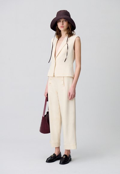 Blazer sans manches beige et pantalon large, assortis à des mocassins noirs et un chapeau seau brun foncé. Tient un sac à bandoulière bordeaux.