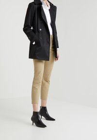 Svart trenchcoat med dubbelknäppt design; krämfärgad skjorta; beige, skräddarsydda byxor; svarta spetsiga ankelstövlar med stilettklack.