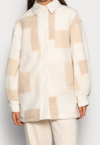Witte fleece oversized button-up shirt met beige blokpatronen, voorzien van een puntige kraag en manchetten voor een textuurafwerking.