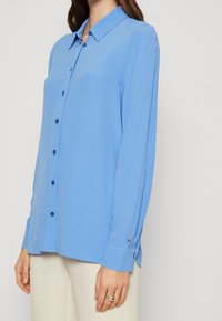 Camicia azzurro chiaro con maniche lunghe, colletto aperto e bottoni blu scuro. Presenta un piccolo etichetta con il logo sul polsino.