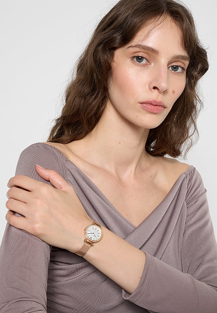 Roségoldenes Mesh-Uhr mit weißem Zifferblatt und blauen Akzenten, am Handgelenk getragen. Der Hintergrund zeigt ein sanft strukturiertes, drapiertes Mauve-Oberteil.