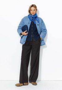 Veste bleue matelassée sur une chemise en denim foncé, associée à un pantalon noir plissé et des chaussures à enfiler imprimé léopard ; une écharpe bleue imprimée complète le look.