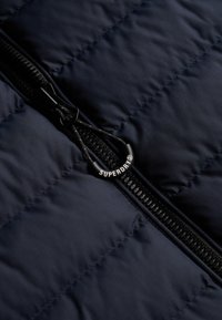Marineblå quiltet jakke med en sort lynlås, som har en mærket pull-tab med teksten "SUPERDRY" i hvid. Blød, glat stofstruktur.