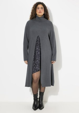 Robe en tricot grise longue avec col montant, présentant une fente avant dévoilant une couche en imprimé léopard violet et noir en dessous. Bottes noires à la cheville.