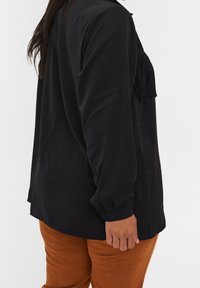 Chemise noire à manches longues avec une poche ornée de franges, associée à un pantalon en velours côtelé couleur rouille. Tissu lisse et léger.