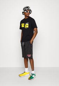 Camiseta negra con estampados gráficos, pantalones cortos a rayas negros, calcetines blancos y zapatillas multicolores; lleva un sombrero de cubo con estampados y gafas de sol.