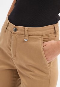 Person in beigen Hosen mit einer Hand in der Tasche und einem schwarzen gerippten Oberteil, das eingesteckt ist, wodurch der Knopf und ein kleines Metallsymbol am Bund sichtbar sind.