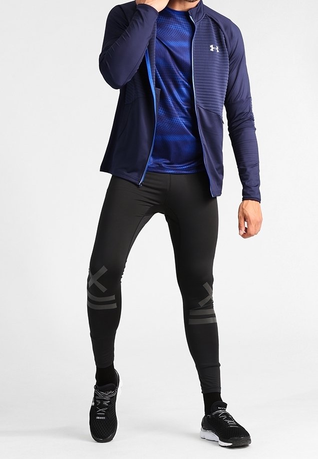 Homme portant une veste de sport zippée bleu marine, une chemise bleue à motifs, des leggings de compression noirs avec des marques grises, et des chaussures de course noires.
