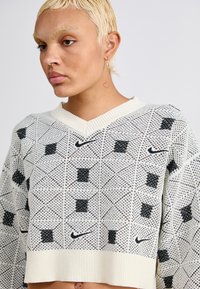 Pārmēru krēmkrāsas džemperis ar melnu ģeometrisku rakstu, V-veida kaklu, ribotām aprocēm un apakšmalu. Priekšpusē ir neliels Nike logo.