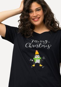 Camiseta negra de cuello en V con un pingüino de dibujos animados vistiendo un gorro amarillo y una bufanda verde, con el texto "Feliz Navidad" arriba.