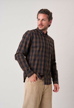 Man die een bruin-zwart geruit overhemd met lange mouwen en beige corduroy broek draagt, kijkt naar links tegen een effen achtergrond.