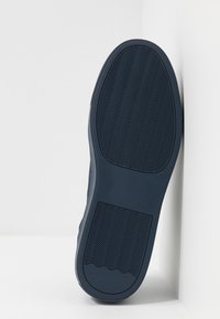 Sulan på en mörkblå sko med vågig mönster, placerad vertikalt mot en vit hörnvägg och golv.