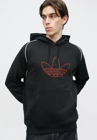 Svart hettegenser med rød omriss-logo, frontlomme og hvite kanter langs skuldrene. Mykt stoff med en strukturert, avslappet passform.