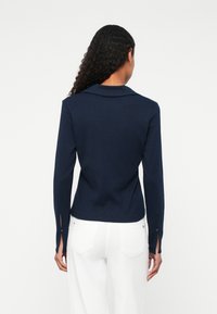 Mørkeblå langermet topp med krage. Har splittede mansjetter med knapper. Glad tekstur, tettsittende design. Brukes med hvite bukser.