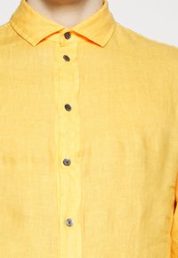 Žlutá lněná košile s špicatým límcem, která má vpředu pět hnědých knoflíků a měkkou, strukturální tkaninu.
