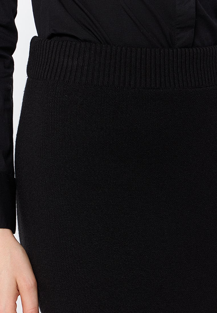 Falda de punto negra con cintura acanalada, que presenta una textura suave y un ajuste entallado, mostrada parcialmente con una camisa negra arriba.
