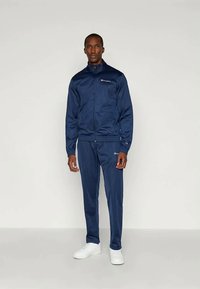 Ναυτικό tracksuit φτιαγμένο από λείο ύφασμα, που περιλαμβάνει ζακέτα με φερμουάρ και παντελόνια με ελαστική μέση, με λογότυπα και στις δύο κομμάτια. Λευκά αθλητικά παπούτσια.