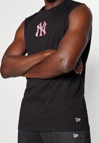 Black sleeveless t-shirt featuring a red and white embroidered logo on the chest. Fabric is soft, with a relaxed fit and crew neckline.