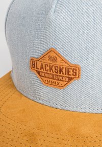 Zilā džinsā ražots cepure ar brūnu zamša vizma. Iezīmēta ar brūnu ādas uzlīmi "BLACKSKIES PREMIUM SUPPLIES" ar iegravētiem detaļām.