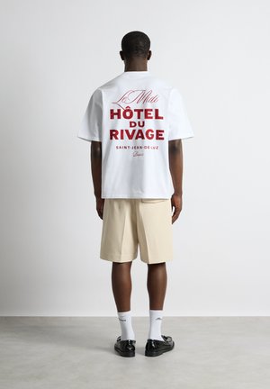 Homme portant un T-shirt blanc avec le texte rouge « Hôtel du Rivage », un short beige, des chaussettes blanches et des chaussures noires, debout face à un mur blanc.
