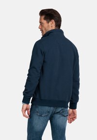 Marineblauwe sweatshirt met ritssluiting en gestructureerde stof, geribbelde manchetten en zoom. Model van achteren afgebeeld, gecombineerd met blauwe jeans.