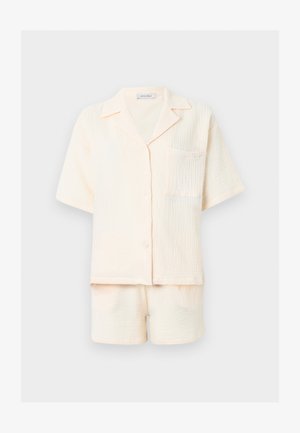 Kreemikujuline tekstuuriga lühikeste varrukatega nööpidega särk rinnataskuga ja sobivate lühikeste pükstega komplekt ühevärvilisel heledal taustal.