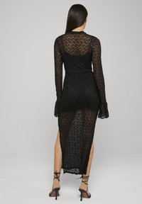 Robe longue à manches longues en dentelle noire avec un design transparent, des fentes latérales et des poignets évasés. La texture est complexe avec un motif floral.