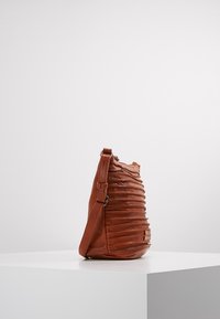 Hnědá kožená taška přes rameno s nařaseným designem, texturovaným povrchem a nastavitelným páskem. Na přední straně má malou značku.