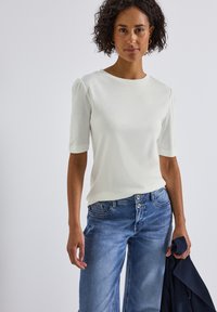 Haut blanc à manches courtes avec épaules bouffantes, associé à un jean en denim bleu clair. Le tissu semble doux avec une texture lisse.