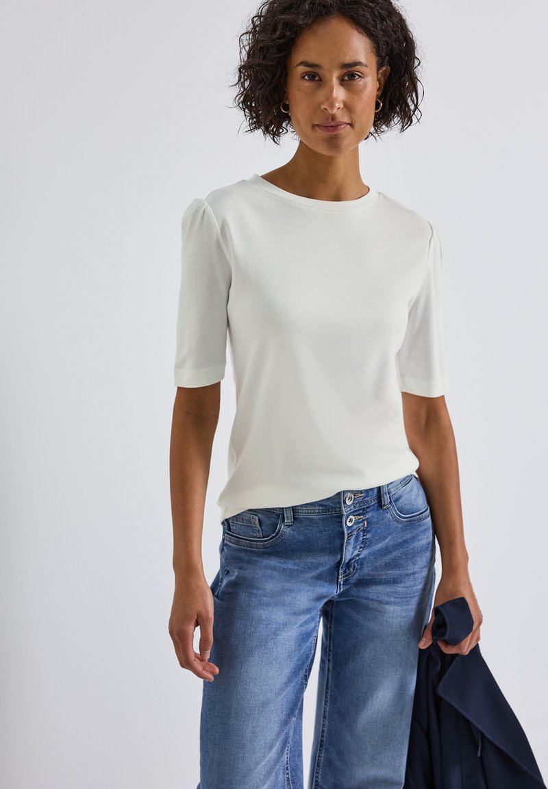 Haut blanc à manches courtes avec épaules bouffantes, associé à un jean en denim bleu clair. Le tissu semble doux avec une texture lisse.